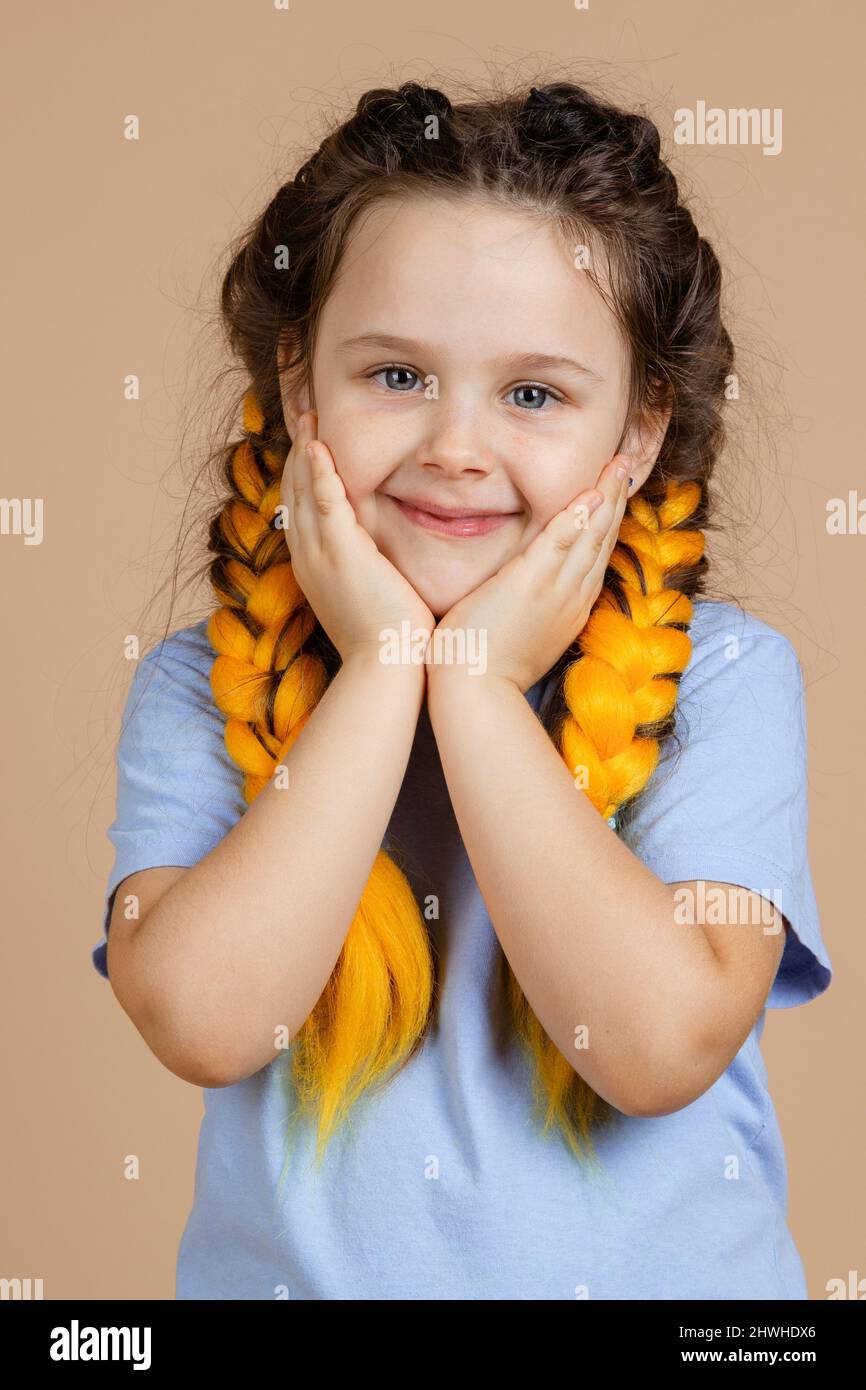 Porträt eines jungen Kindes mit Händen auf den Wangen leicht lächelnd Blick auf die Kamera mit leuchtenden Augen mit gelben Kanekalon Zöpfe auf dem Kopf auf beige Stockfoto