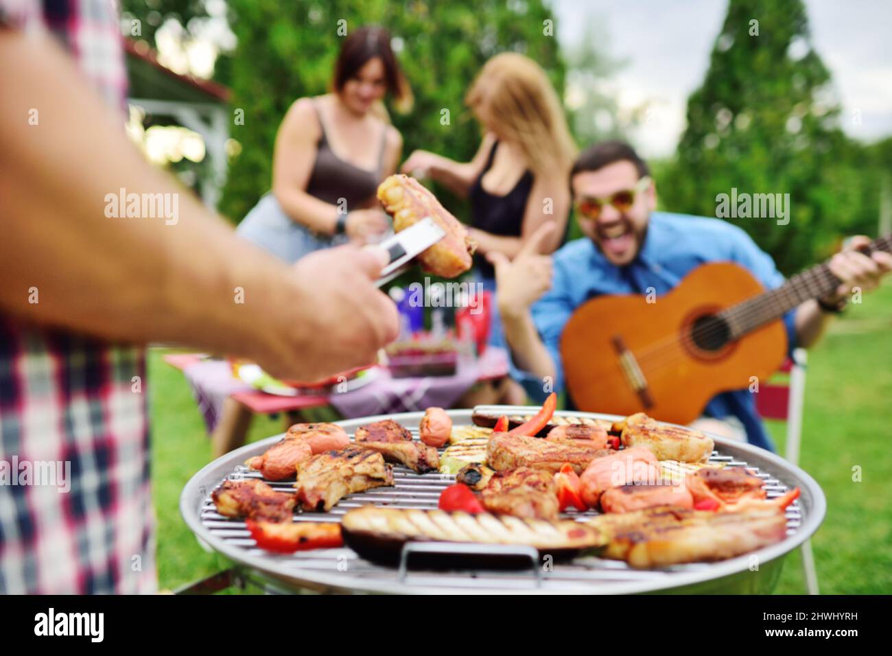 Die Hand des Mannes hält eine Grillzange mit einem saftig leckeren Fleischsteak vor dem Hintergrund eines Grillgrills mit Fleisch und Gemüse und einer Gruppe von Stockfoto