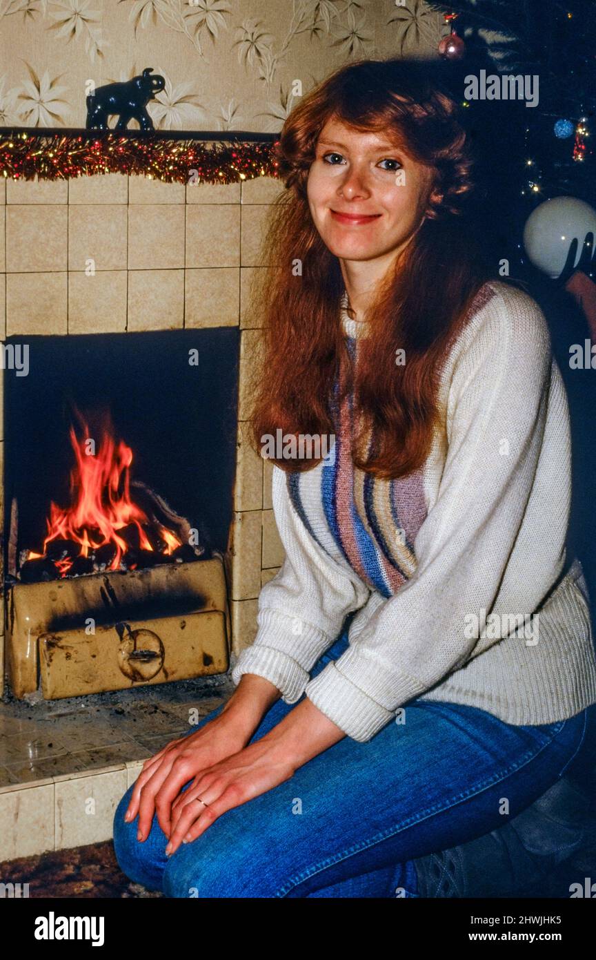 Lächelnde junge Frau mit roten Haaren, die sich durch ein offenes Kohlefeuer warm hält 1980s Rugby england Stockfoto