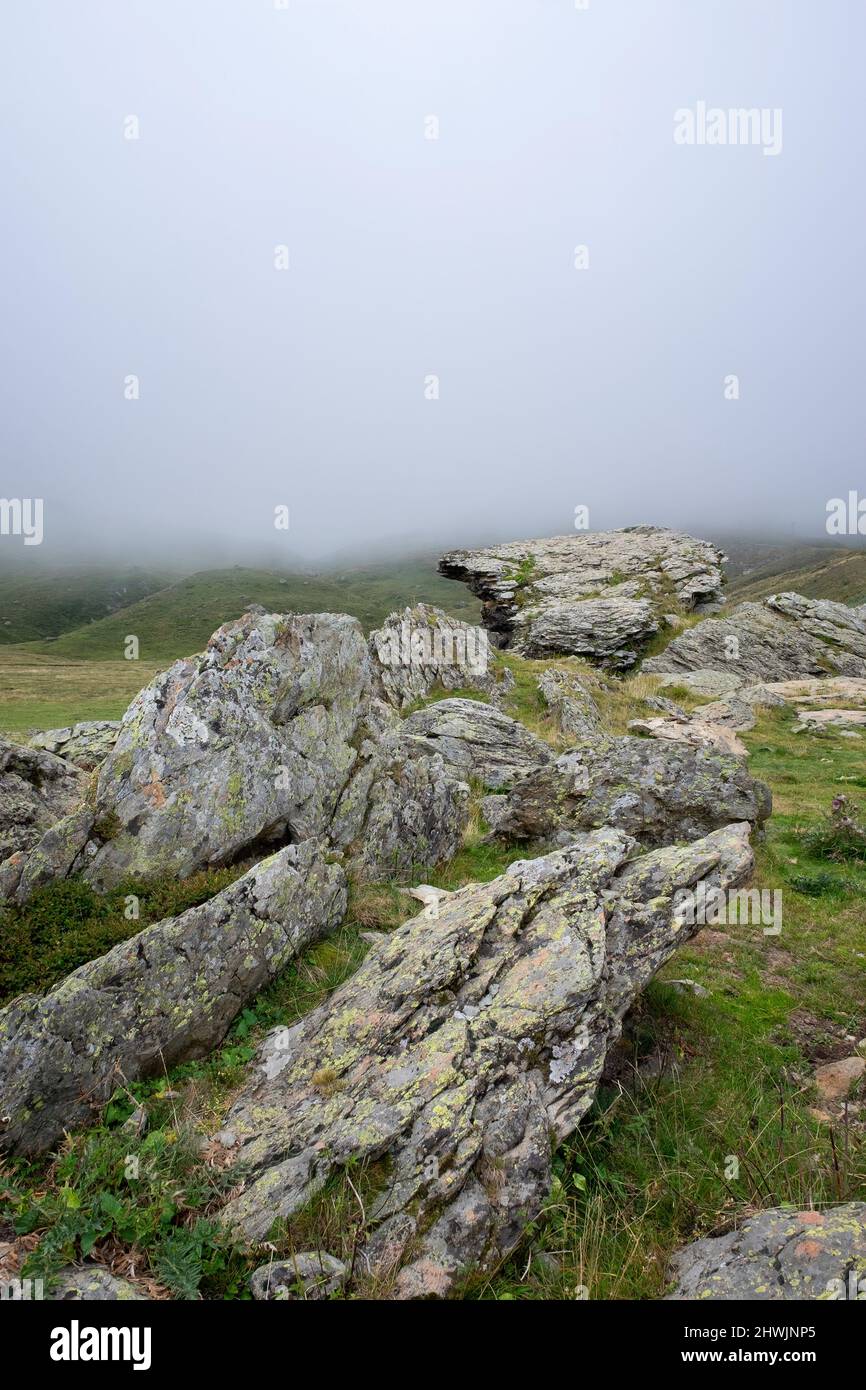 Hochgebirgslandschaft mit granitischen Felsen mit Flechten im Vordergrund und einer dicken Nebelbank, die die Berge im Hintergrund bedeckt, senkrecht Stockfoto