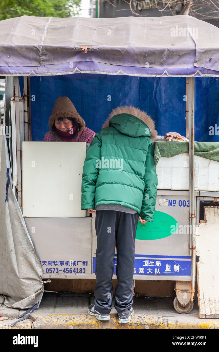 Kunde im grünen Mantel, der nach dem Angebot eines schnellen Imbissbudes auf der Straße fragt. Jiashan, China Stockfoto