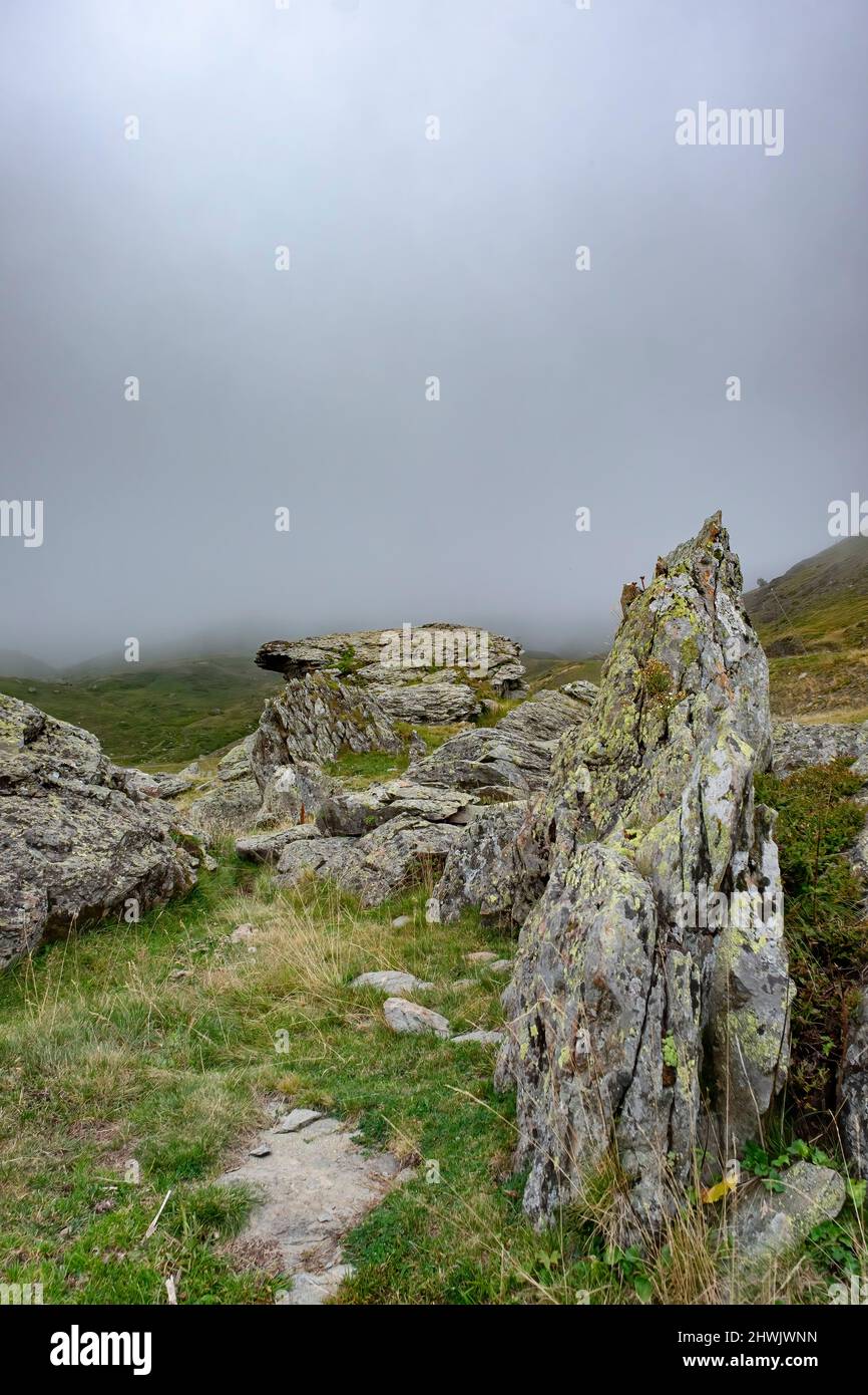 Hochgebirgslandschaft mit granitischen Felsen, die sich im Vordergrund mit Flechten bedecken, und einer dicken Nebelbank, die die Berge im Hintergrund bedeckt, verti Stockfoto