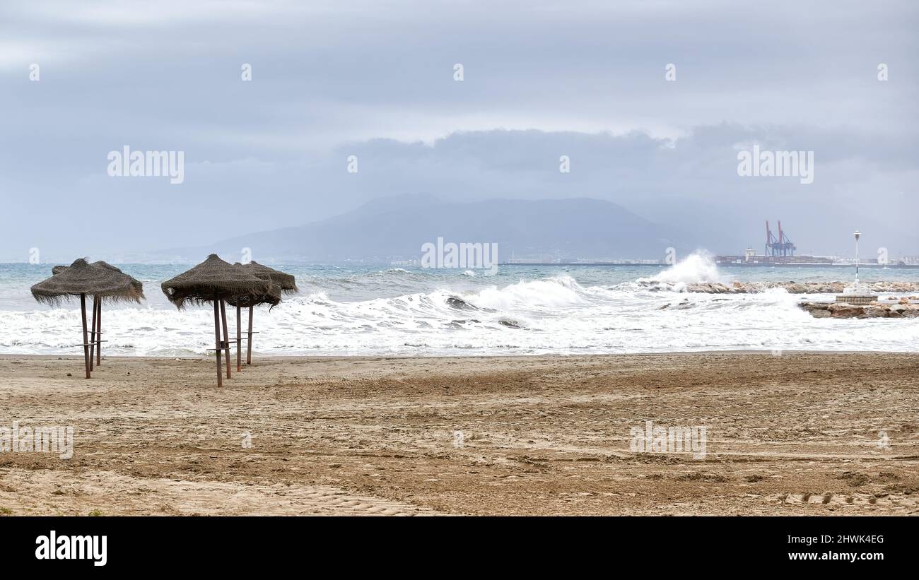 Schönes Bild des Strandes in der Stadt Malaga, Spanien im Winter mit leeren Sonnenschirmen, starken Wellen, die gegen das Wellenbrecher und den por krachen Stockfoto