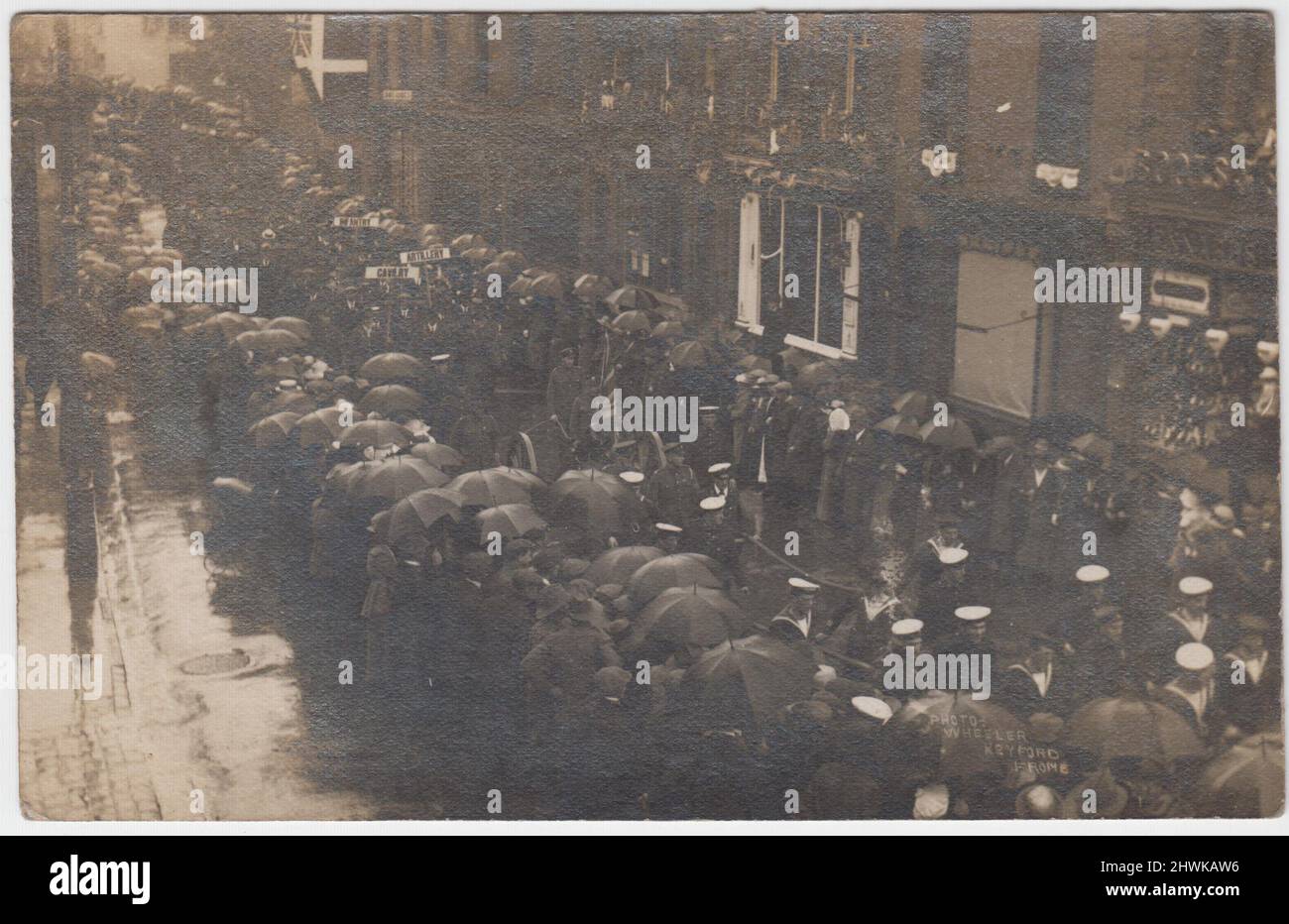Parade von Männern, die während des Ersten Weltkriegs in Keyford, Frome, einmarschieren, um sich zu vermarken. Die Parade wird von Matrosen und Soldaten in Uniform angeführt, gefolgt von Männern in Zivilkleidung, die Schilder mit den Aufschrift „Kavallerie“, „Artillerie“ und „Infanterie“ tragen. Die Straße ist von Zivilisten gesäumt, die Regenschirme halten, um den starken Regen zu verhindern Stockfoto