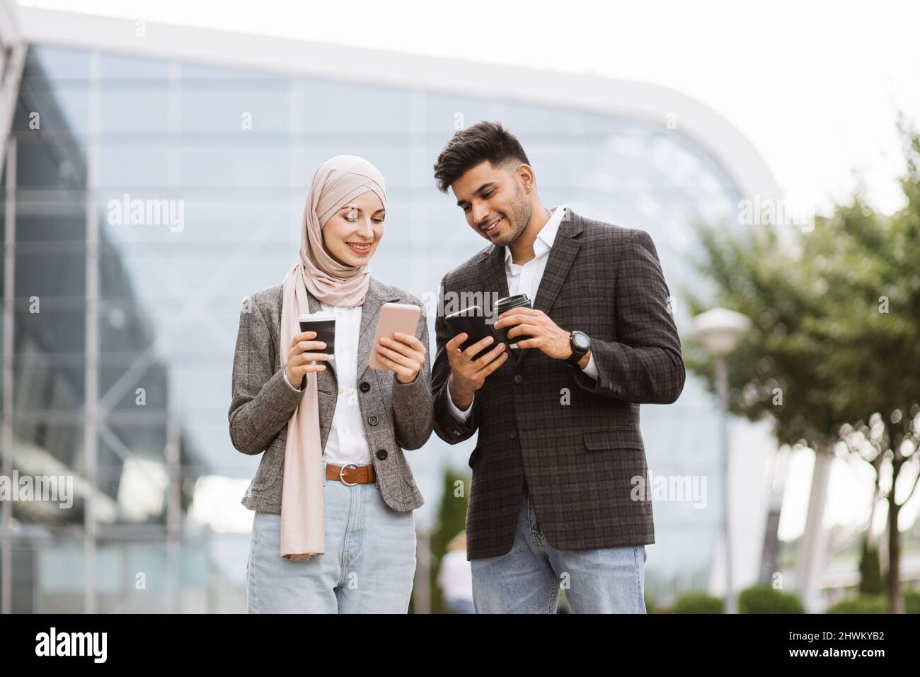 Arabisches Geschäftspaar, das Spaß hat und sich während der Kaffeepause auf der Straße unterhält. Frau in Hijab zeigt ihrem Kollegen ihren Smartphone-Bildschirm und teilt Ideen oder Nachrichten Stockfoto