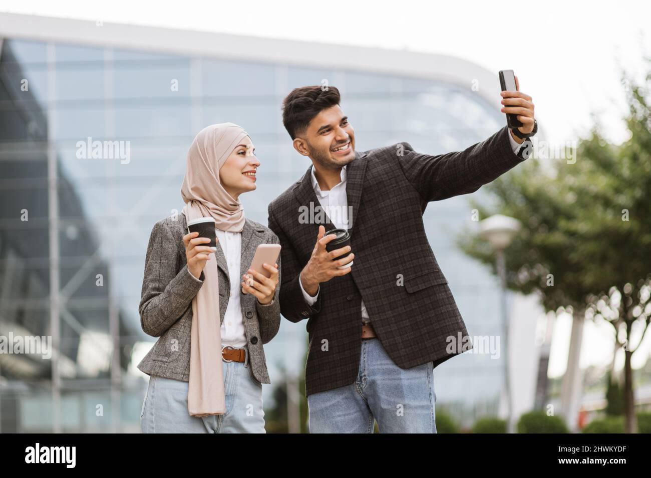 Geschäftsleute bei einer Kaffeepause im Freien. Glücklicher, sympathischer hindu-Mann, der mit seinem Bürokollegen spricht, hübsche muslimische Frau in Hijab und Selfie auf dem Smartphone macht. Gemeinsam Kaffee trinken Stockfoto