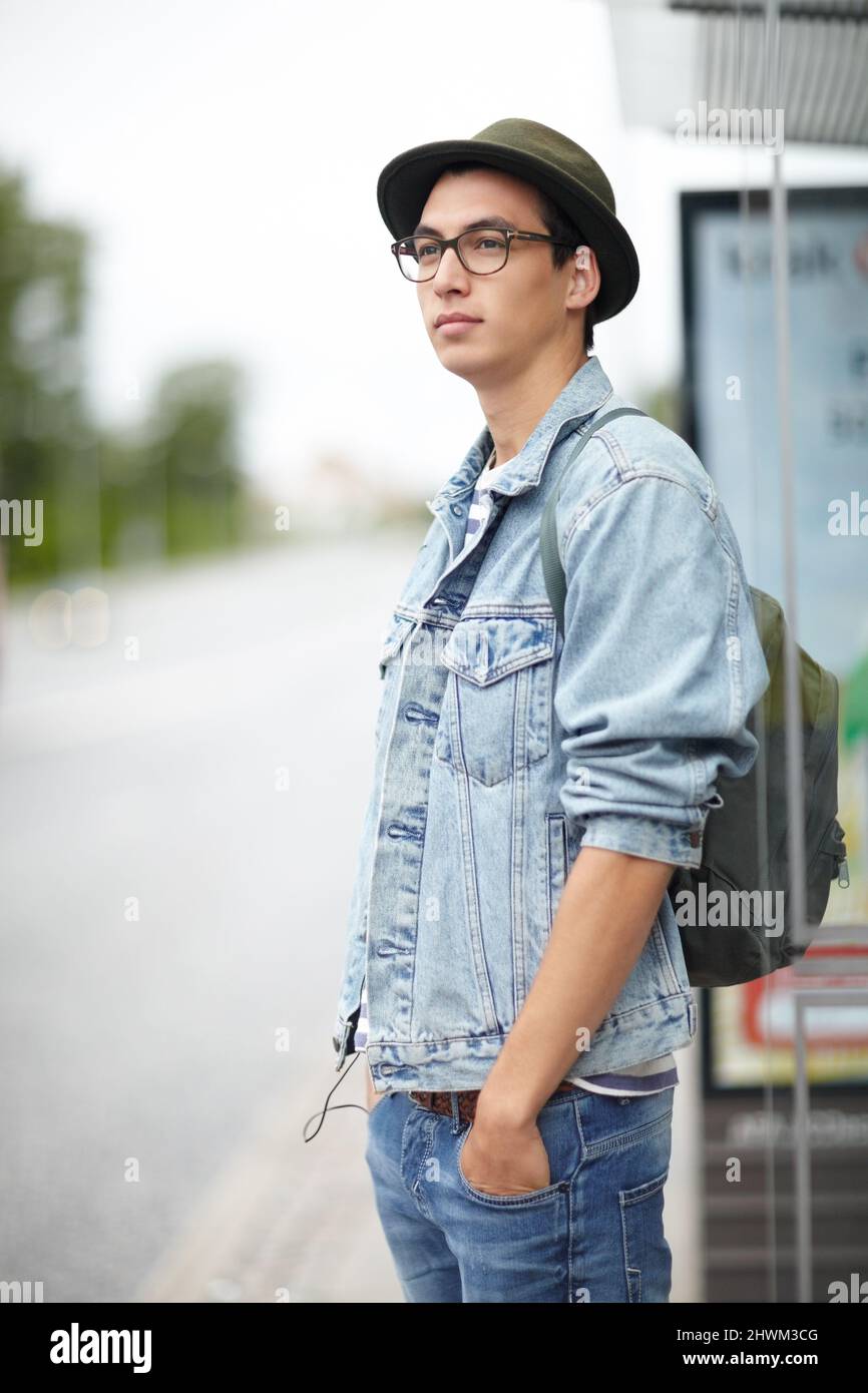 Warten entlang der Busroute. Angesagter junger Mann, der an einer Bushaltestelle steht, mit den Händen in den Taschen. Stockfoto