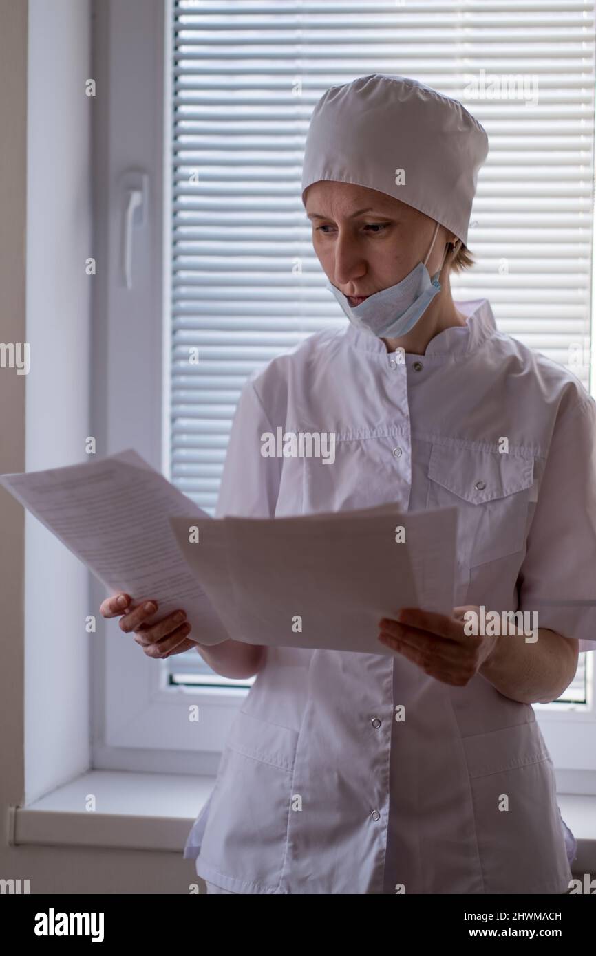 Eine Ärztin mit einer Maske auf ihrem Gesicht steht neben dem Fenster und schaut durch die Dokumente. Stockfoto