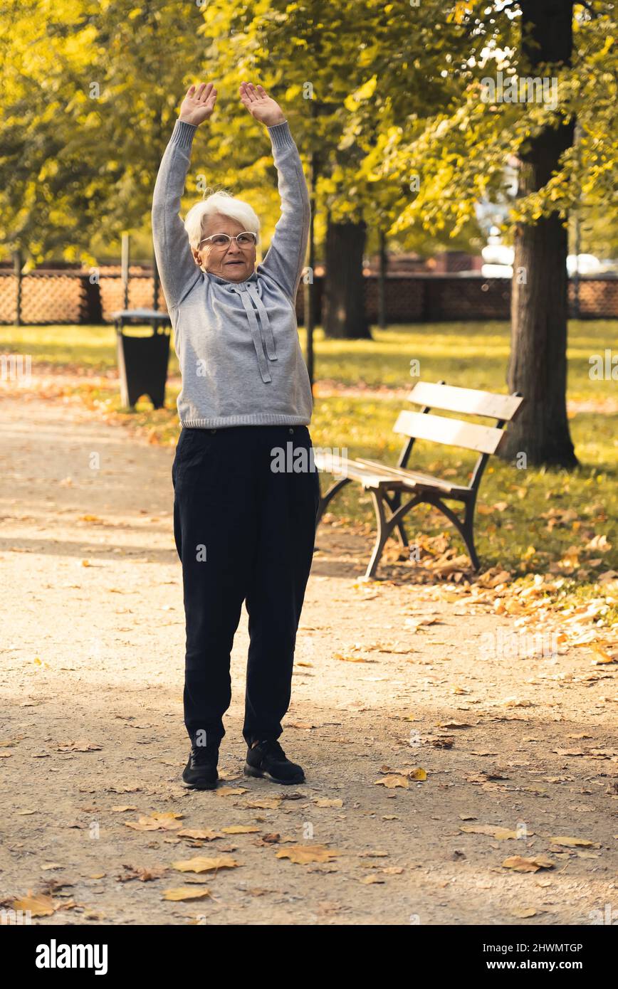 Vertikale Aufnahme einer älteren, im Ruhestand gezogenen grauhaarigen Frau, die ihre Arme in einem öffentlichen Park streckte und dabei die frische Luft und das wunderschöne Herbstwetter genoss. Hochwertige Fotos Stockfoto