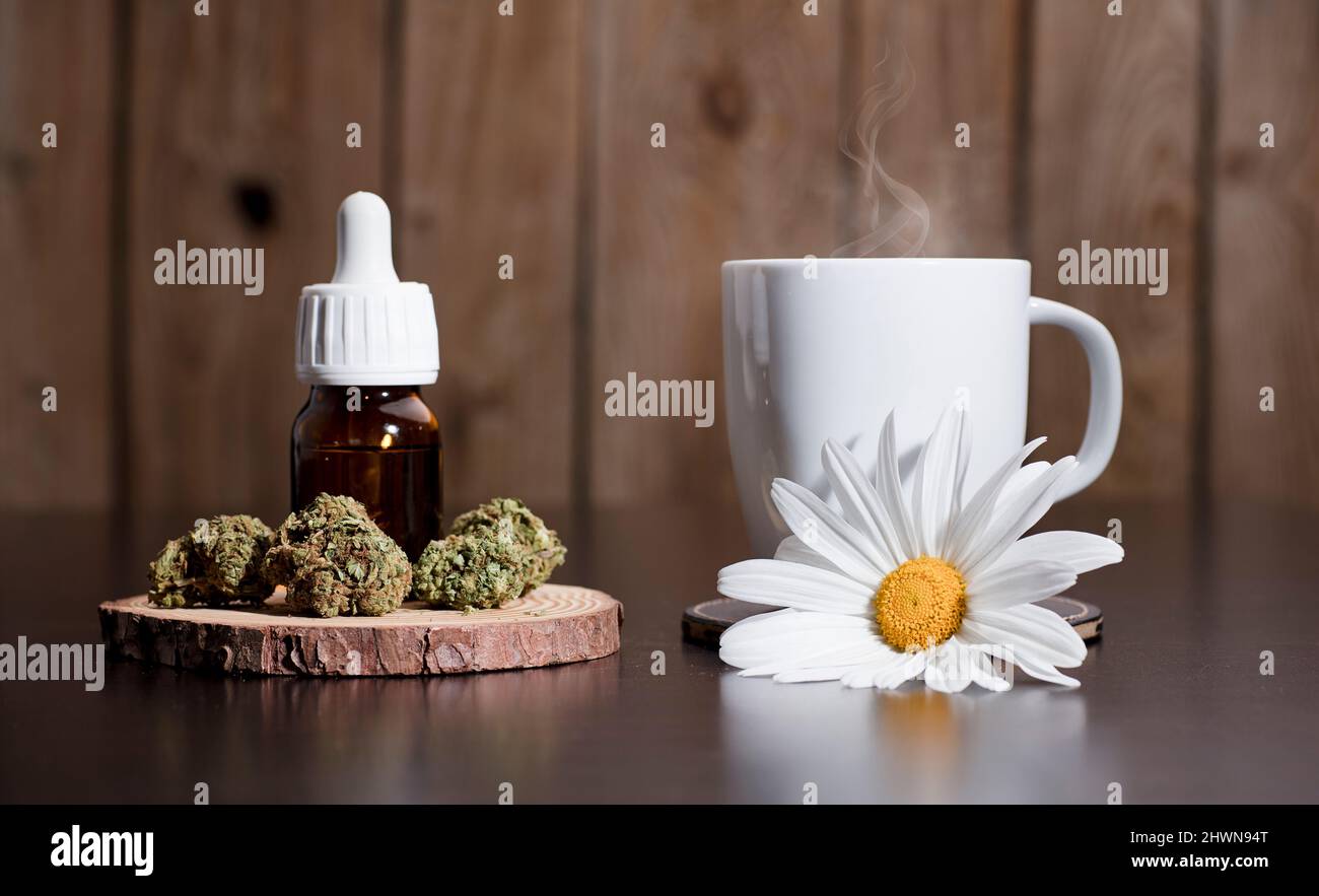 Marihuana Knospen mit Tropfen Spender Flasche, weiße Tasse mit Dampf Infusion Gänseblümchen Blume Ornament, auf braunem Tisch, Holzhintergrund Stockfoto