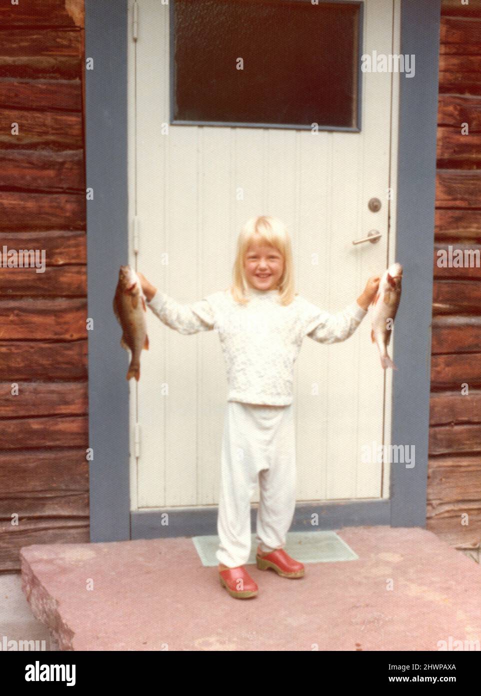 Originalfoto der 1970er Jahre von einem jungen blonden Mädchen, das zwei Fische vor einer Kabine in Schweden hält Stockfoto