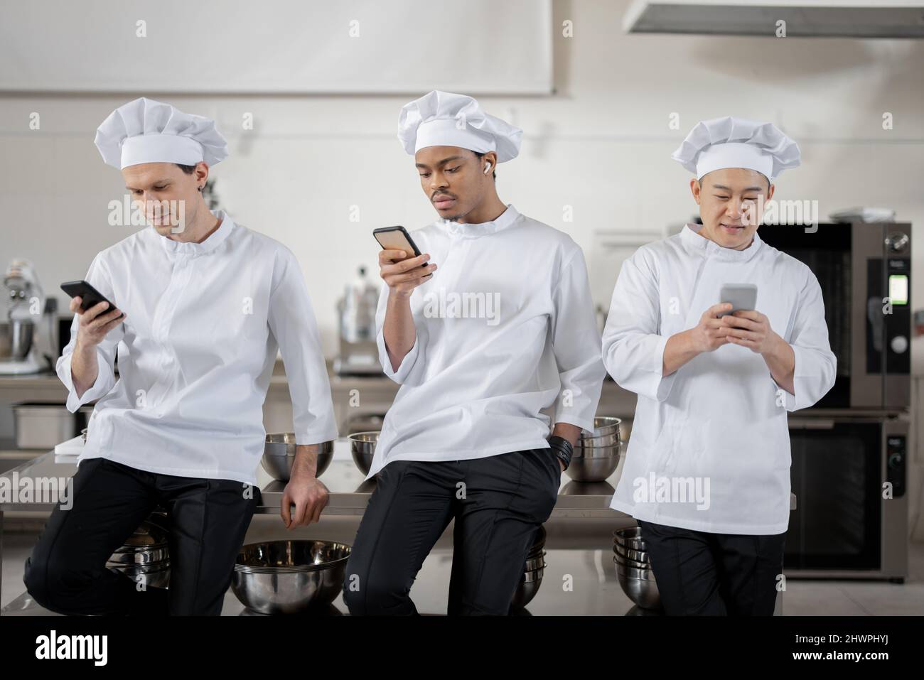 Drei multirassische Köche stehen während einer Pause in der Restaurantküche mit Smartphones. Asiatische, lateinische und europäische Männer in weißer Uniform während einer Arbeitspause Stockfoto