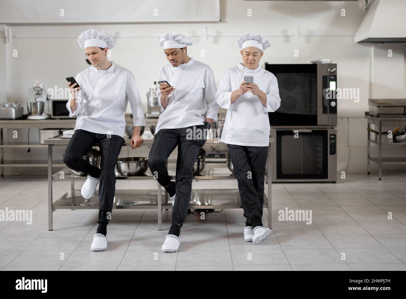 Drei multirassische Köche stehen während einer Pause in der Restaurantküche mit Smartphones. Asiatische, lateinische und europäische Männer in weißer Uniform während einer Arbeitspause Stockfoto