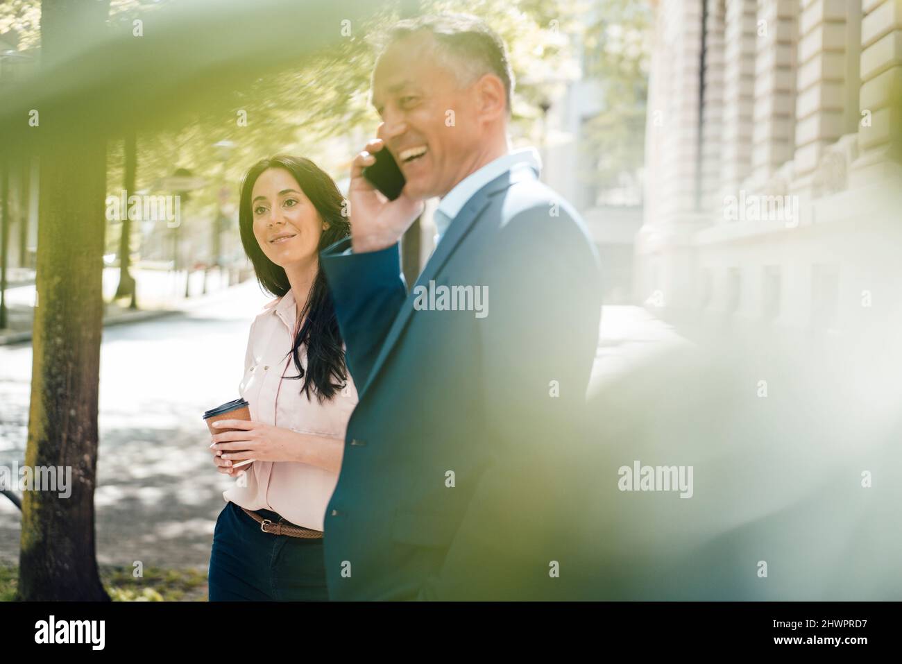 Fröhlicher Geschäftsmann, der auf dem Mobiltelefon von einer Geschäftsfrau auf dem Fußweg spricht Stockfoto