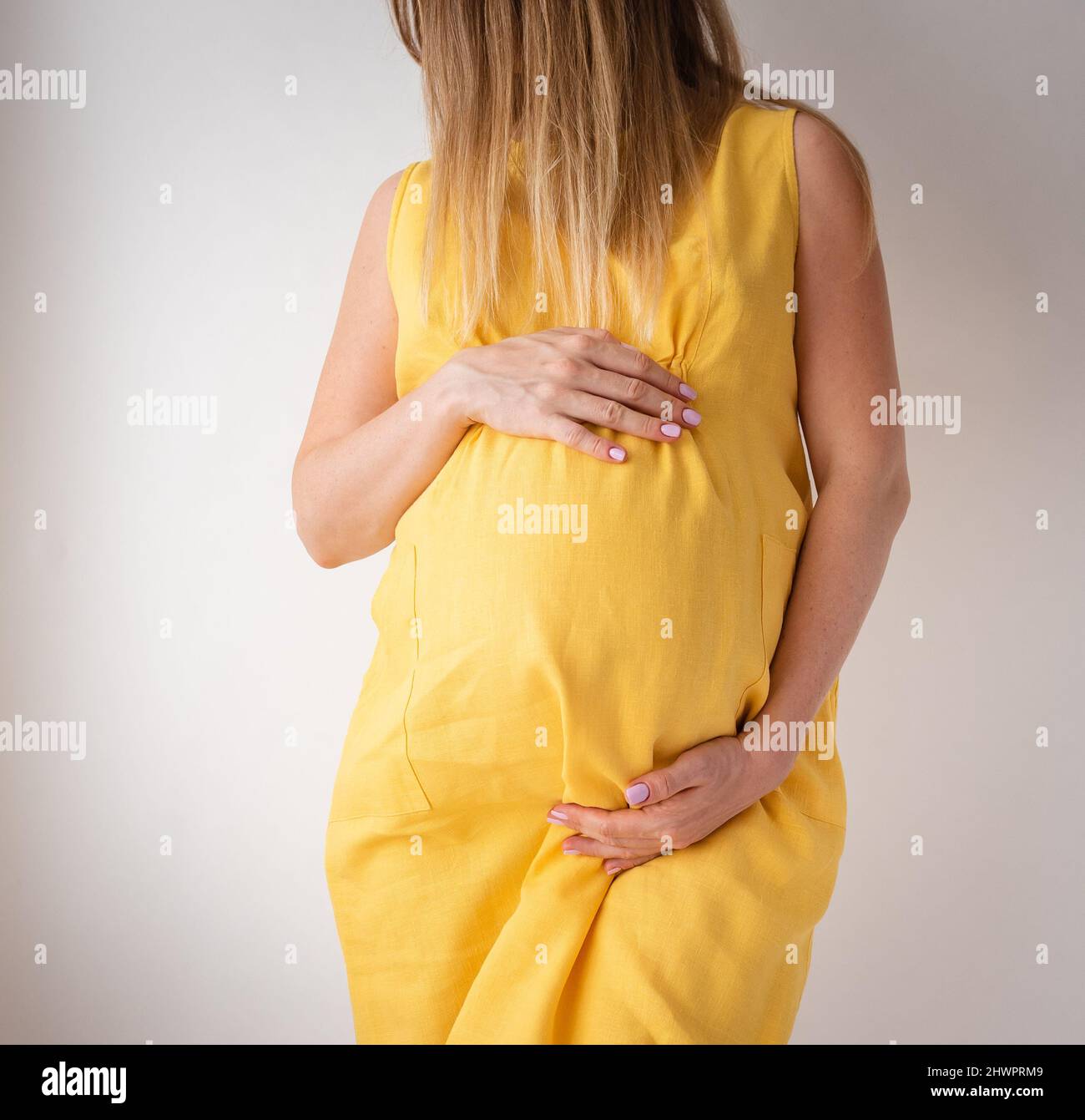 Die Schwangere in einem gelben Kleid steht auf weißem Hintergrund mit den Händen auf dem Bauch, dem Konzept der Weiblichkeit Stockfoto