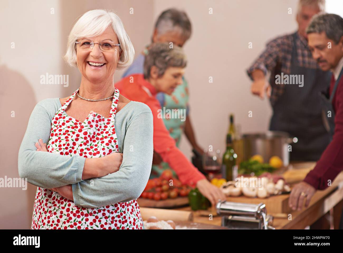 Ich liebe es, Dinner-Partys zu werfen. Porträt einer älteren Frau mit Freunden, die im Hintergrund zusammen kochen. Stockfoto