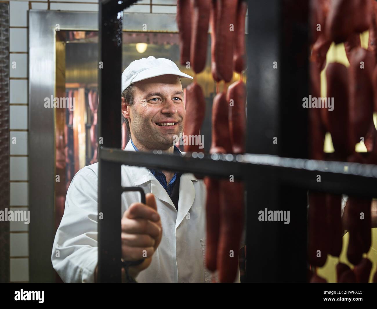 Lächelnder Metzger zieht Würstchen aus dem Rauchhaus bei der Metzgerei Stockfoto