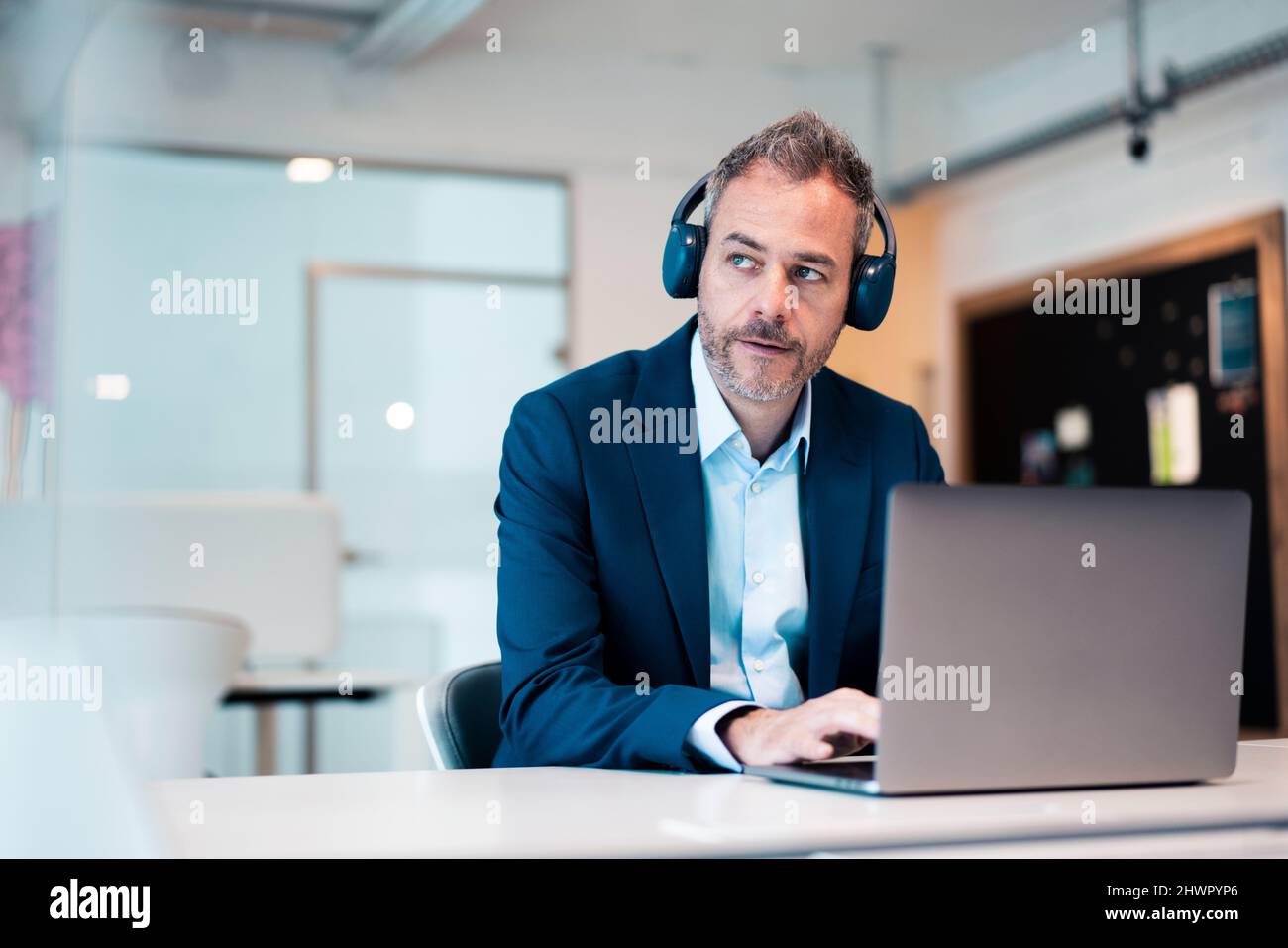 Ein nachdenklicher Geschäftsmann mit kabellosen Kopfhörern und Laptop am Arbeitsplatz Stockfoto