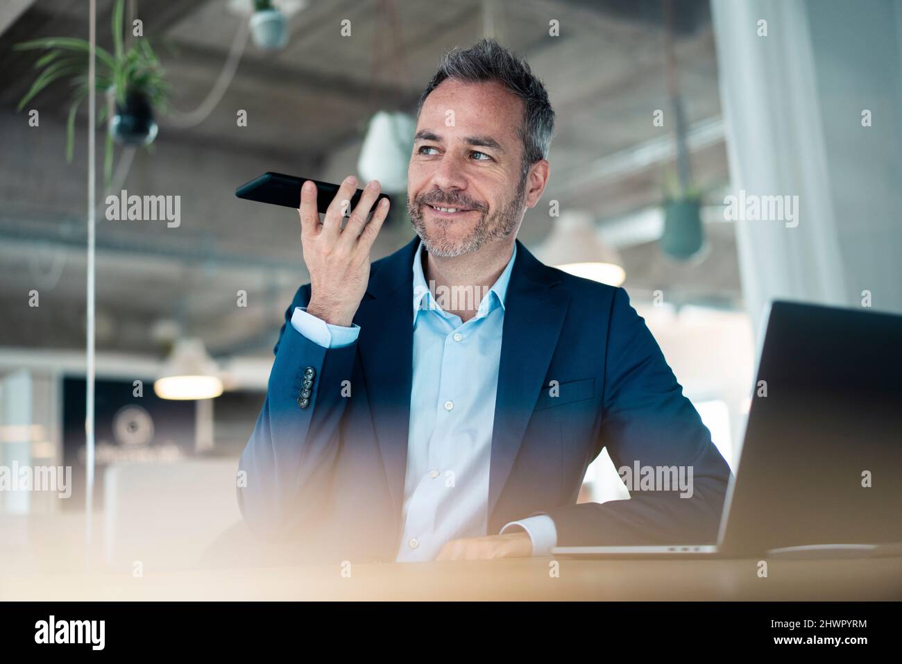 Lächelnder Geschäftsmann, der am Arbeitsplatz mit einem Laptop auf dem Mobiltelefon sitzt Stockfoto