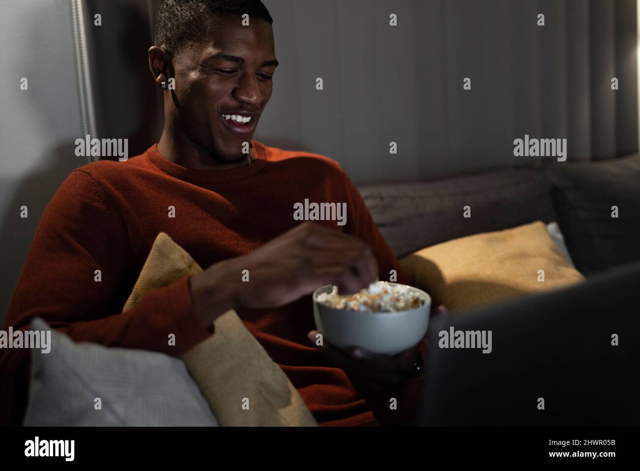 Lächelnder Mann, der sich einen Film auf dem Laptop ansieht und zu Hause Popcorn isst Stockfoto