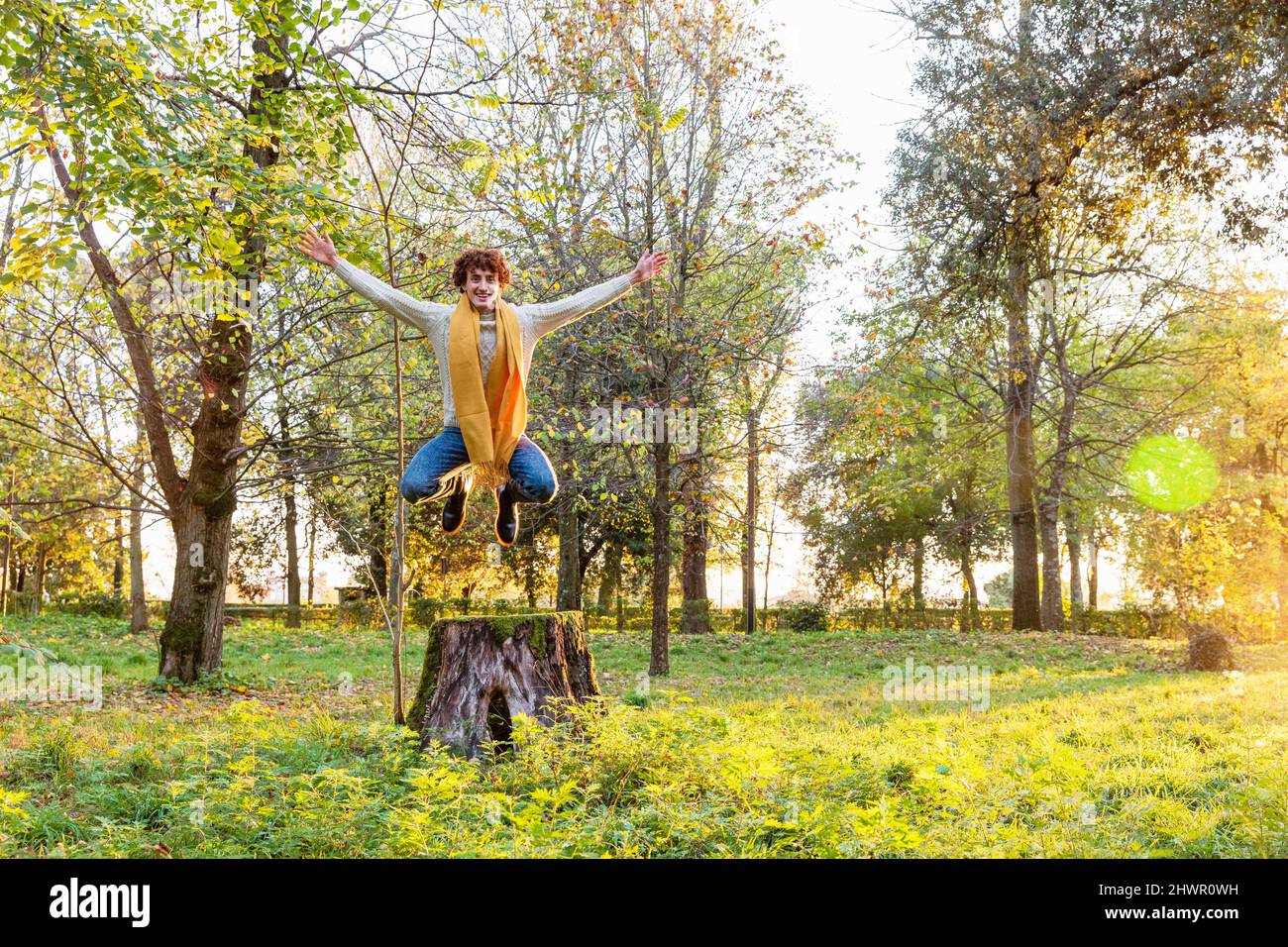 Junger Mann, der im Herbstpark mit ausgestreckten Armen aus dem Baumstumpf springt Stockfoto
