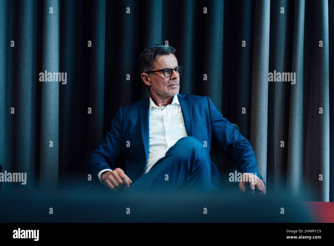 Kontemplativer Geschäftsmann, der im Büro vor dem Vorhang auf einem Stuhl sitzt Stockfoto