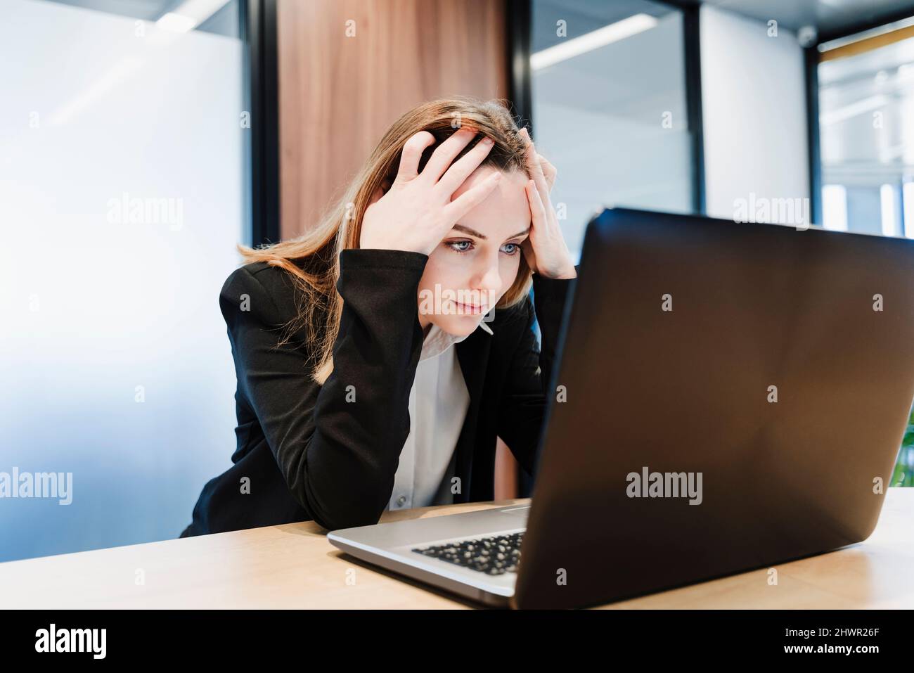 Frustrierte Geschäftsfrau mit Kopf in den Händen und Blick auf den Laptop im Büro Stockfoto