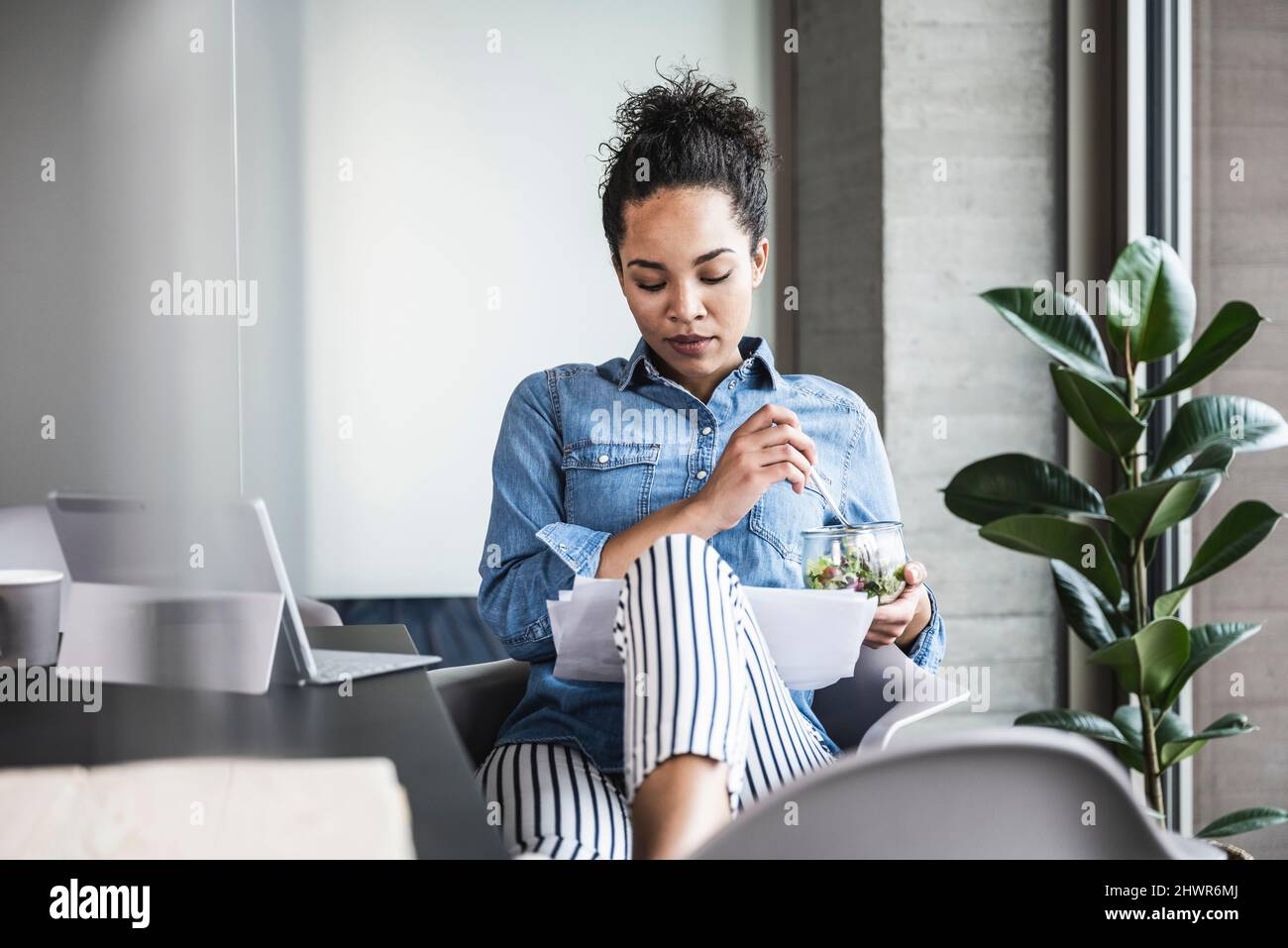 Junge Geschäftsfrau mit Lebensmittelschale, die Dokumente am Arbeitsplatz analysiert Stockfoto