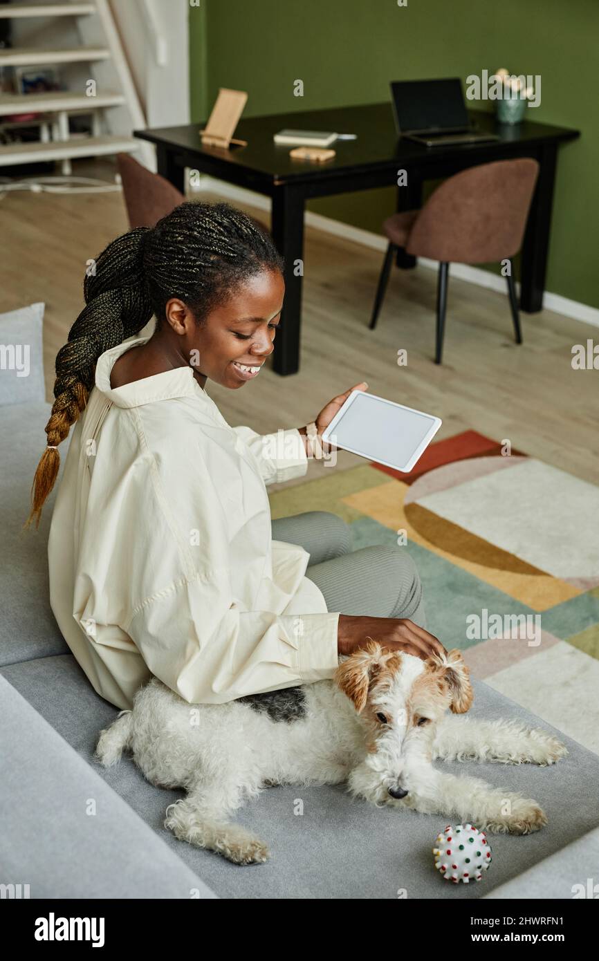 Vertikales Porträt einer lächelnden schwarzen Frau, die den Hund streichelt, während sie sich zu Hause auf der Couch entspannt Stockfoto