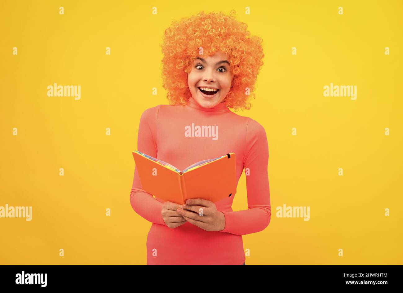 Glückliches Mädchen Kind mit orangen Haaren in rosa Poloneck Lächeln hält Schulbuch gelben Hintergrund, Bildung. Stockfoto