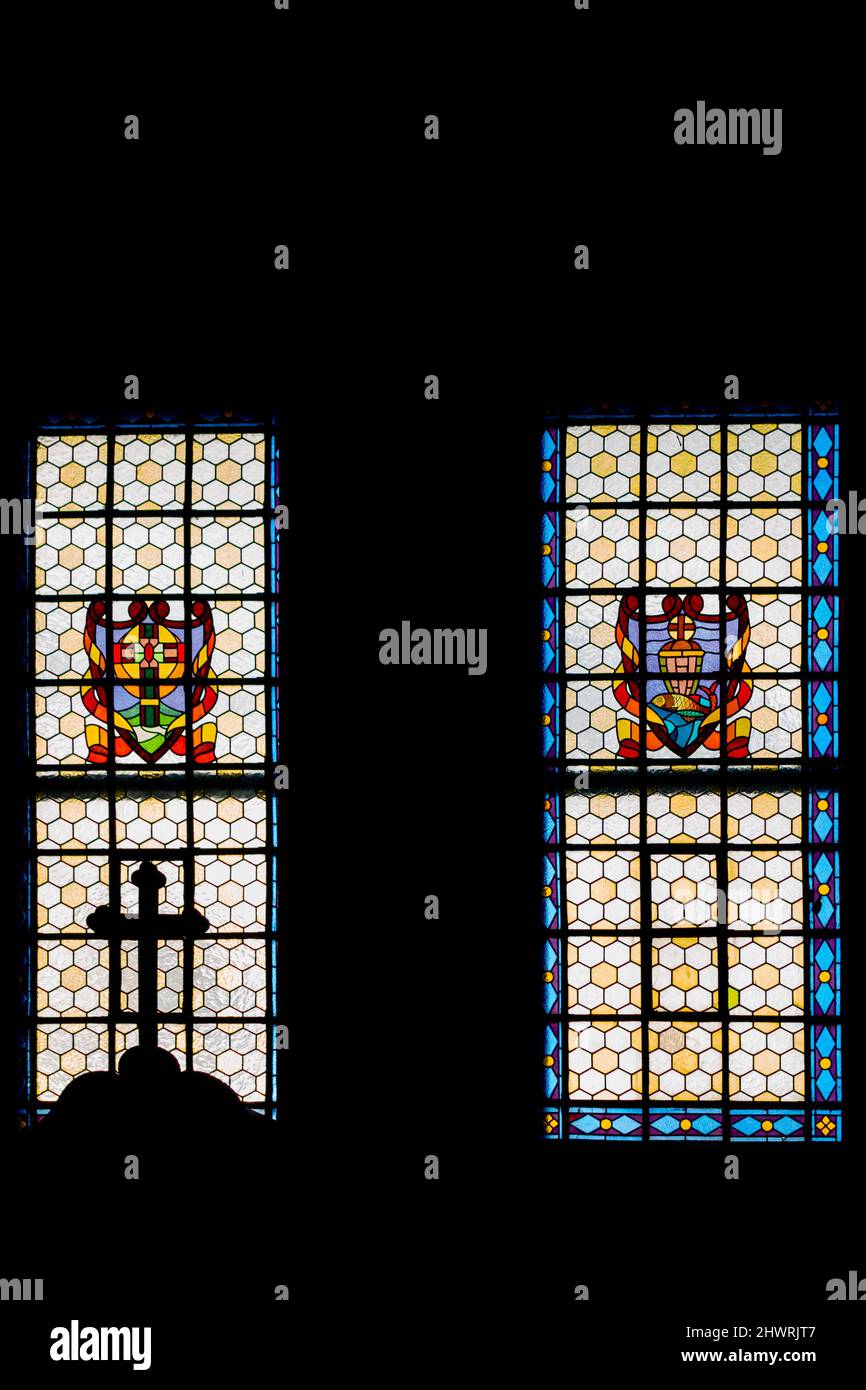 Das katholische Kreuz auf der Spitze des Beichtstuhls vor dem Hintergrund eines sonnendurchfluteten Buntglasfensters. Das Foto wurde in der Kirche aufgenommen. Stockfoto