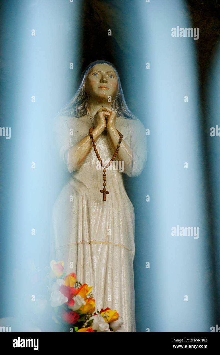 Die betende Jungfrau Maria-Statue in der Grotte Notre Dame des Sees in der Nähe des Sees Annecy, durch verschwommene Zaunbars gesehen. Duingt, Haute-Savoie, Frankreich. Stockfoto