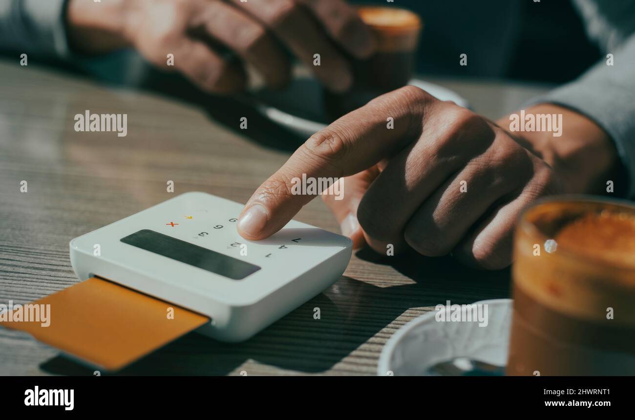 Nahaufnahme eines jungen Mannes, der an einem Tisch eines Straßencafés sitzt und den PIN-Code seiner Kreditkarte in das Yepad eines drahtlosen Zahlungsterminals einfügt Stockfoto