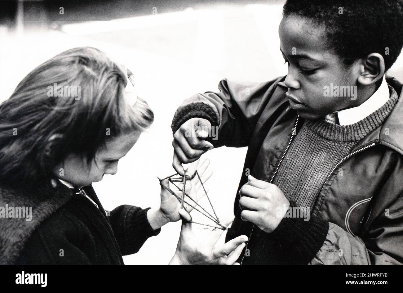 Ein junges Mädchen und ein Junge spielen ein altmodisches Spiel namens Cat's Cradle. Wiki beschreibt es als ein Spiel, bei dem verschiedene Saitenfiguren zwischen den Fingern erzeugt werden, entweder einzeln oder durch Übergeben einer Schnur hin und her. Um 1975 in einem Schulhof in Brooklyn, New York. Stockfoto