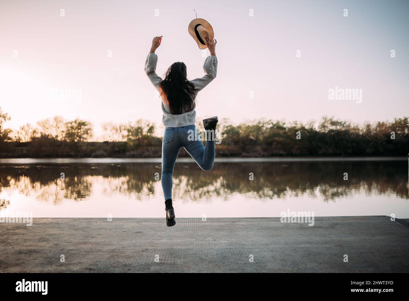 Unerkennbare Brünette Frau, die vor Freude am Ufer eines Flusses mit einem Hut in der Hand springt Stockfoto