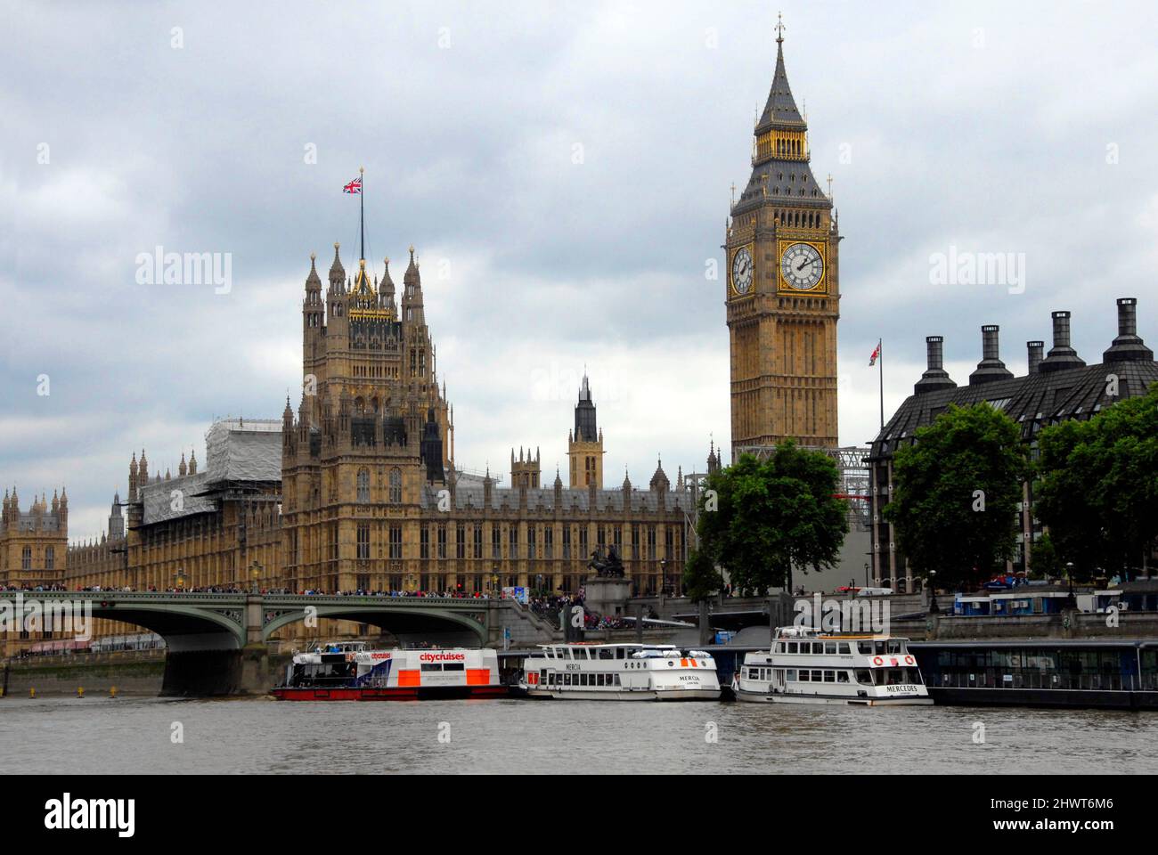 Houses of Parliament, London mit einem Teil der Westminster Bridge und touristischen Vergnügungsbooten, die bei typisch bewölktem Wetter vor Anker liegen Stockfoto