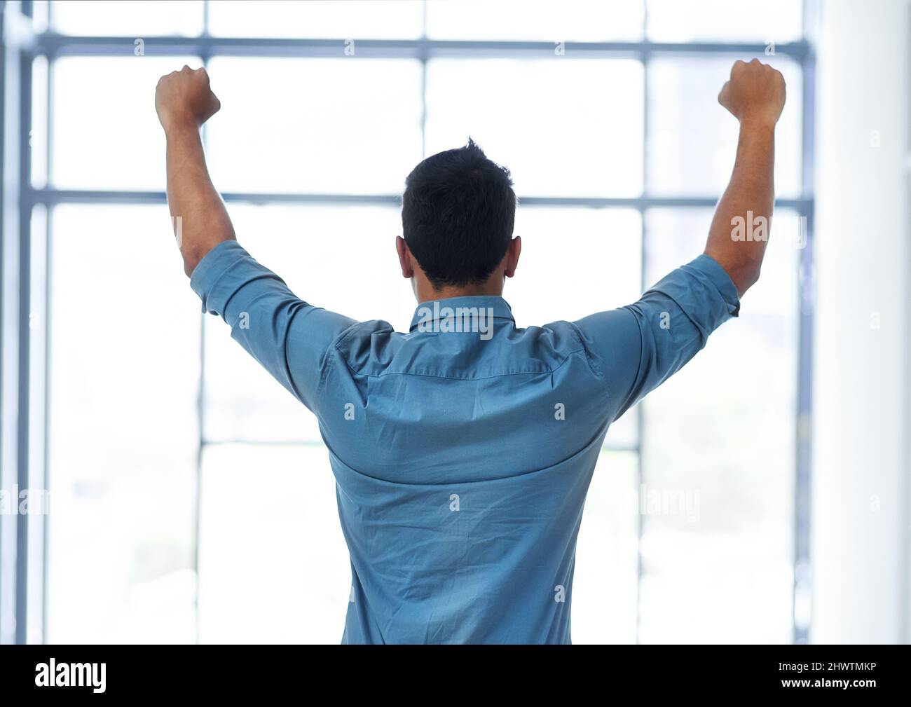 Ich habe es noch einmal getan. Rückansicht eines nicht erkennbaren Geschäftsmannes, der in einem modernen Büro mit erhobenem Arm feiert. Stockfoto