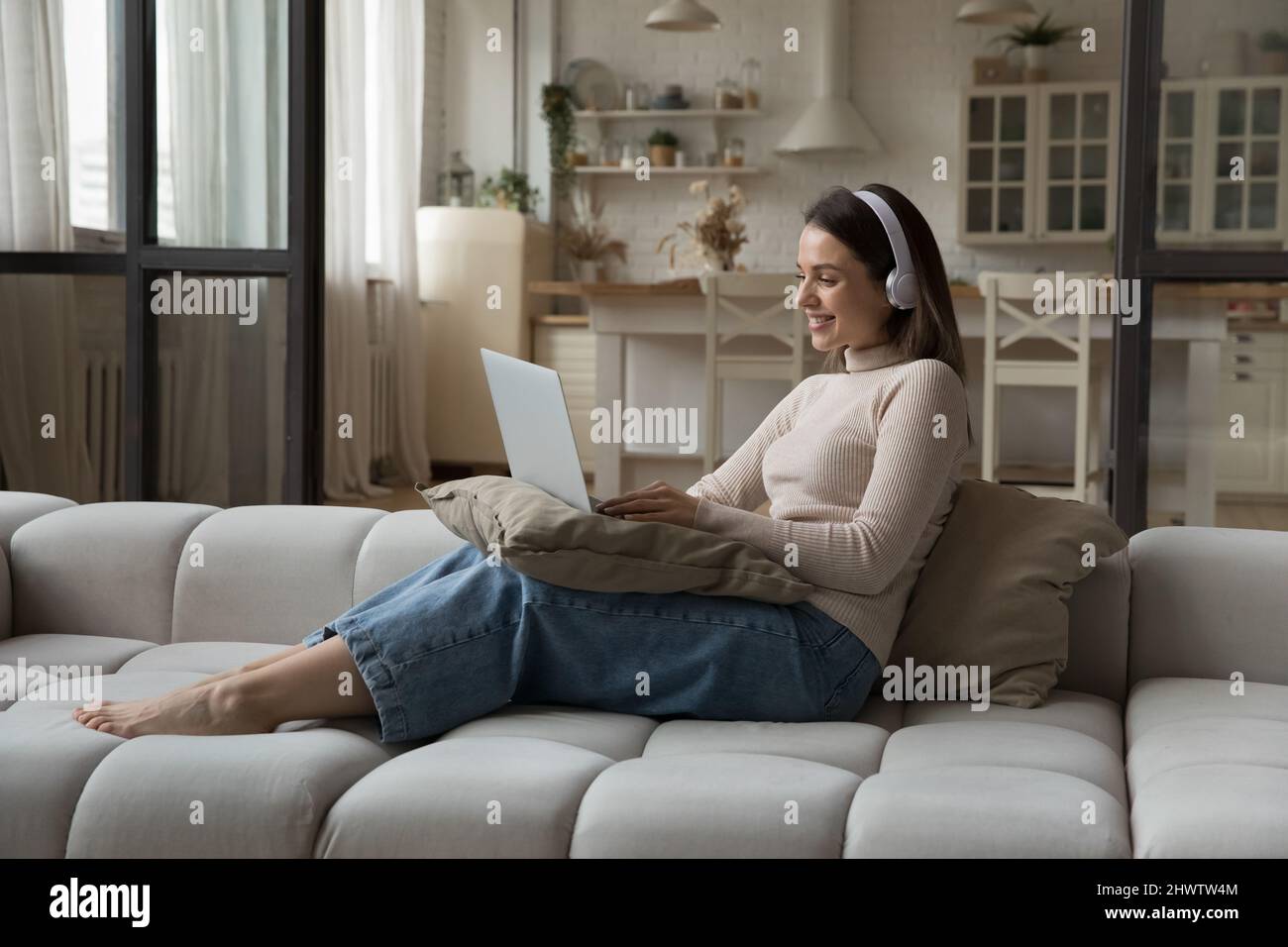 Lächelnde junge Frau mit Kopfhörern benutzt einen Laptop, der auf dem Sofa sitzt Stockfoto