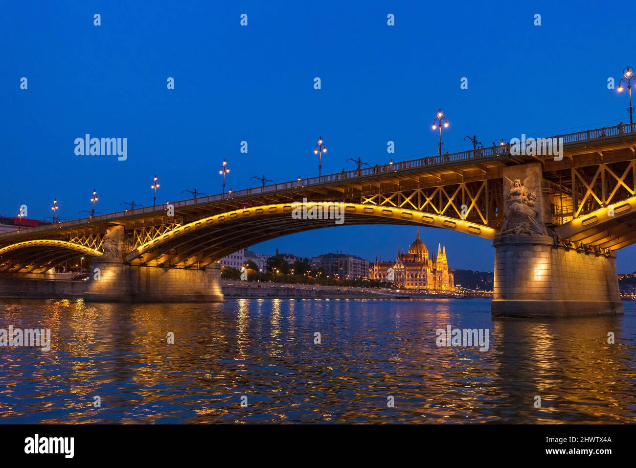 Die Margaretenbrücke mit dem Parlamentsgebäude im Hintergrund in Budapest, Ungarn, beleuchtet über der Donau bei Nacht. Stockfoto