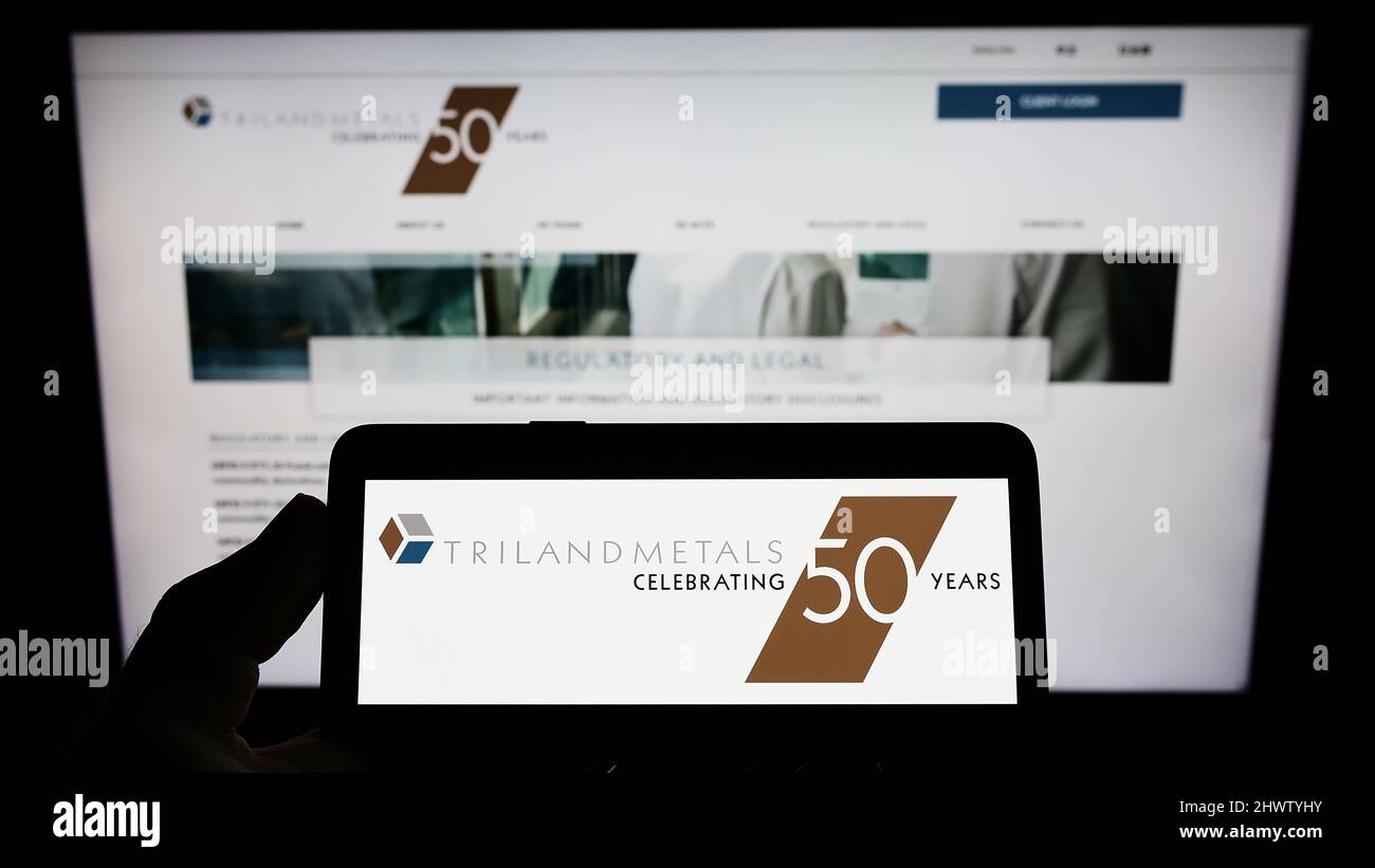 Person mit Mobiltelefon und Logo des britischen Unternehmens Triland Metals Limited auf dem Bildschirm vor der Business-Webseite. Konzentrieren Sie sich auf die Telefonanzeige. Stockfoto