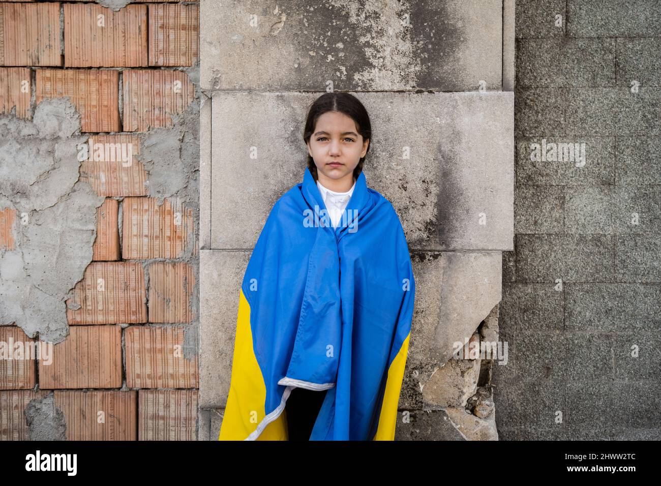 Kleines Mädchen mit ukrainischer Flagge vor einer durch Bomben zerstörten Mauer. Das kleine Mädchen schwenkt die Nationalflagge und betet für den Frieden Stockfoto