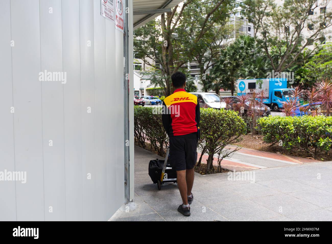 Ein Mann in DHL-Uniform kommt zur Auslieferung, indem er einen tragbaren Trolley mit 2 Rädern im zentralen Geschäftsviertel von Singapur schiebt. Stockfoto