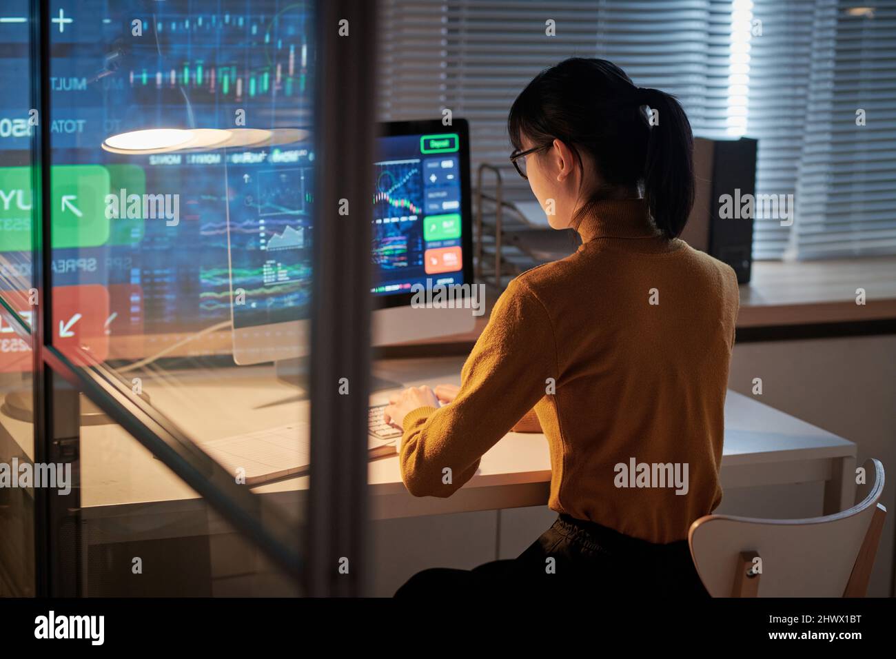 Rückansicht einer jungen Frau, die am Tisch vor dem Computermonitor saß und bis spät in die Nacht mit Software im Büro arbeitete Stockfoto