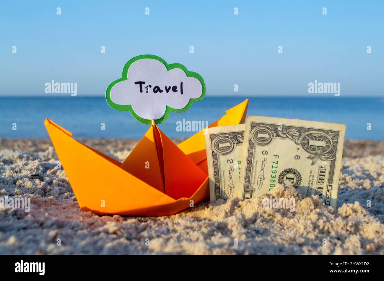 Zwei Papierrechnungen mit einem Dollar, die zur Hälfte in Sand und orangefarbenem Papierboot vergraben sind Stockfoto