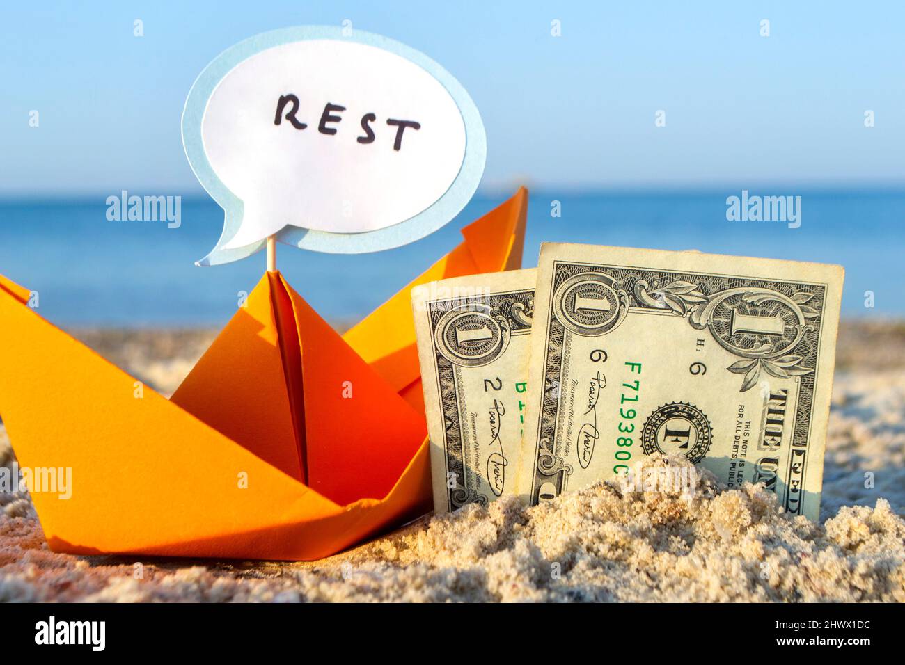 Zwei Papierrechnungen mit einem Dollar, die zur Hälfte in Sand und orangefarbenem Papierboot vergraben sind Stockfoto