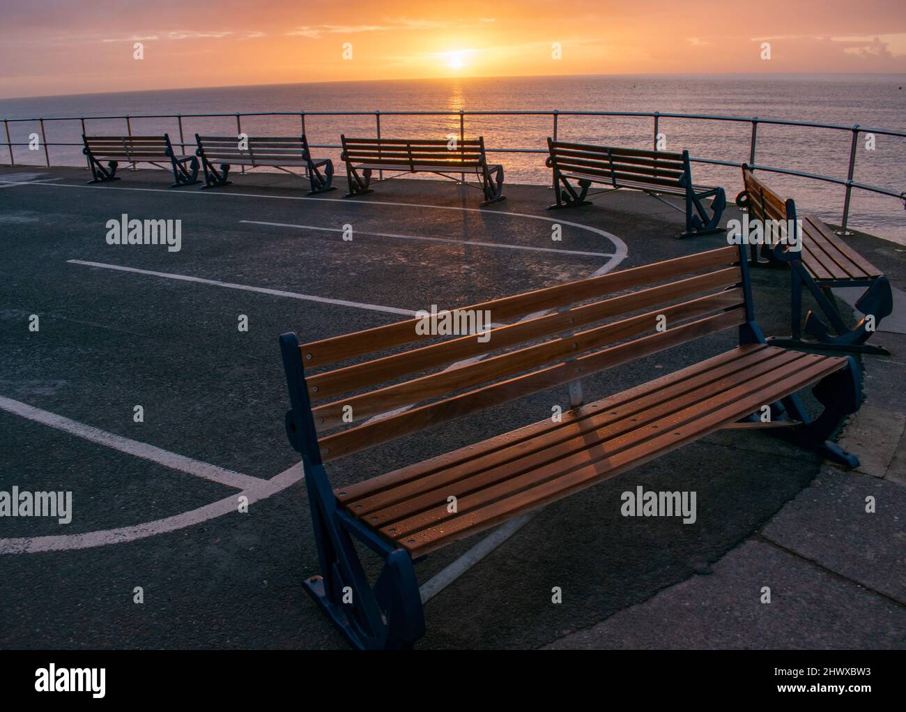Eine geschwungene Reihe von sechs Holzbänken zeigt an einem kalten, klaren Morgen auf das Meer. Bernsteinfarbenes Sonnenlicht bringt sie in ein orangefarbenes Leuchten, während sie leer stehen Stockfoto
