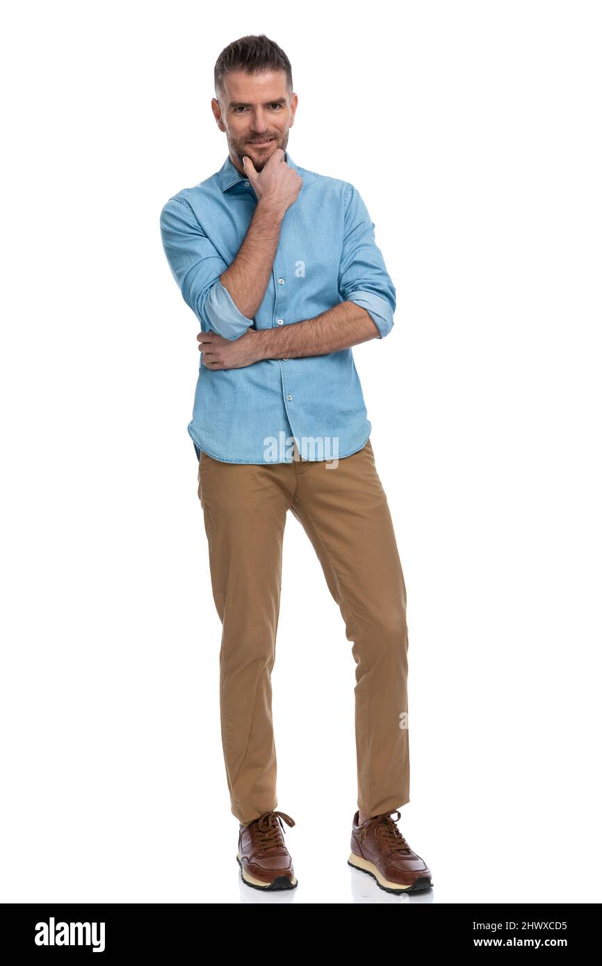 Ganzkörperbild eines jungen Casual-Mannes im Denim-Hemd, der sich über die Arme kreuzt, die Hand zum Kinn hält und denkt und im Studio auf weißem Hintergrund kreativ ist Stockfoto