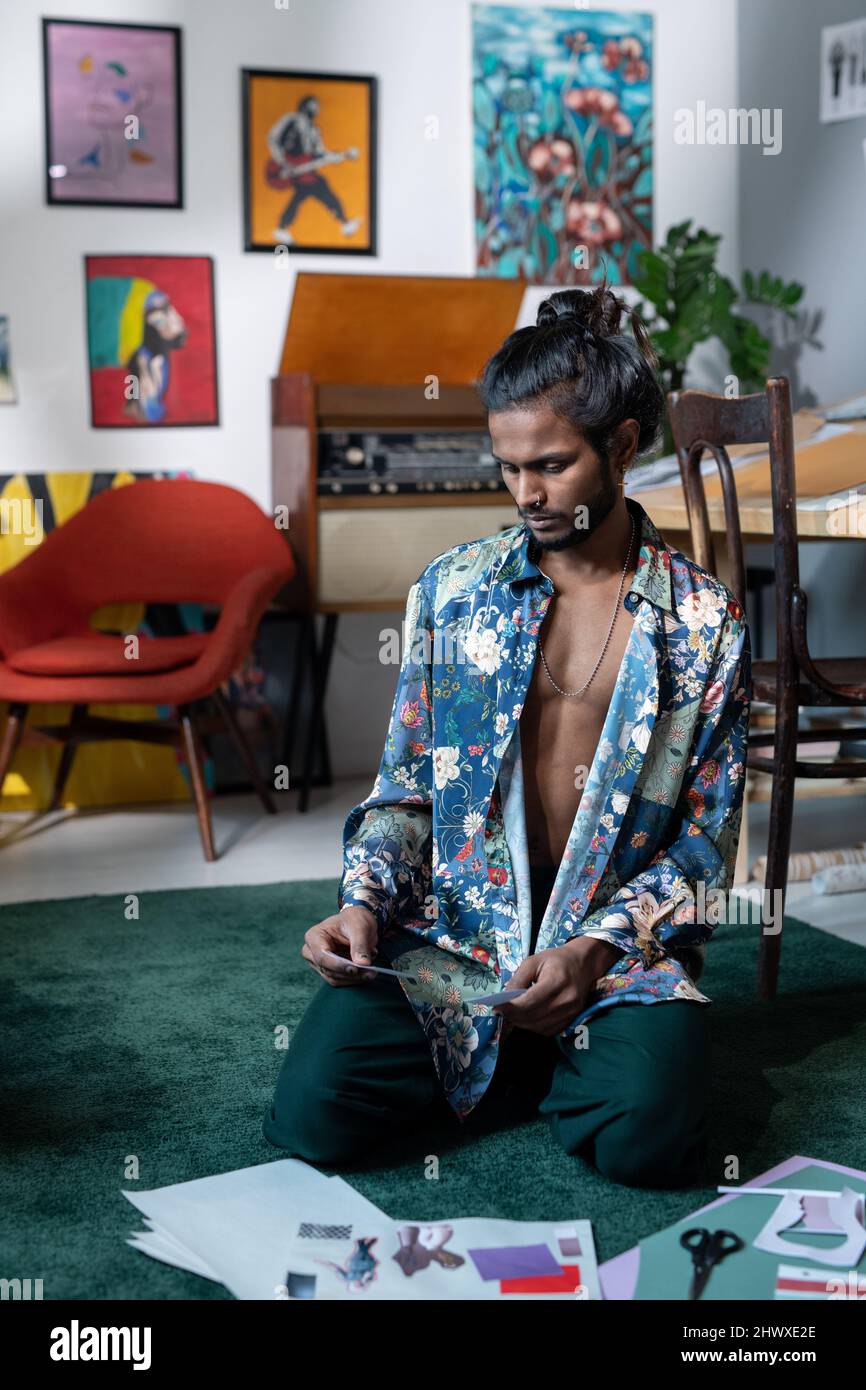 Kreativer Mann im modischen Hemd, der Fotos für kreative Collagen auswählt, während er im Wohnzimmer oder im Kunststudio auf dem Boden sitzt Stockfoto