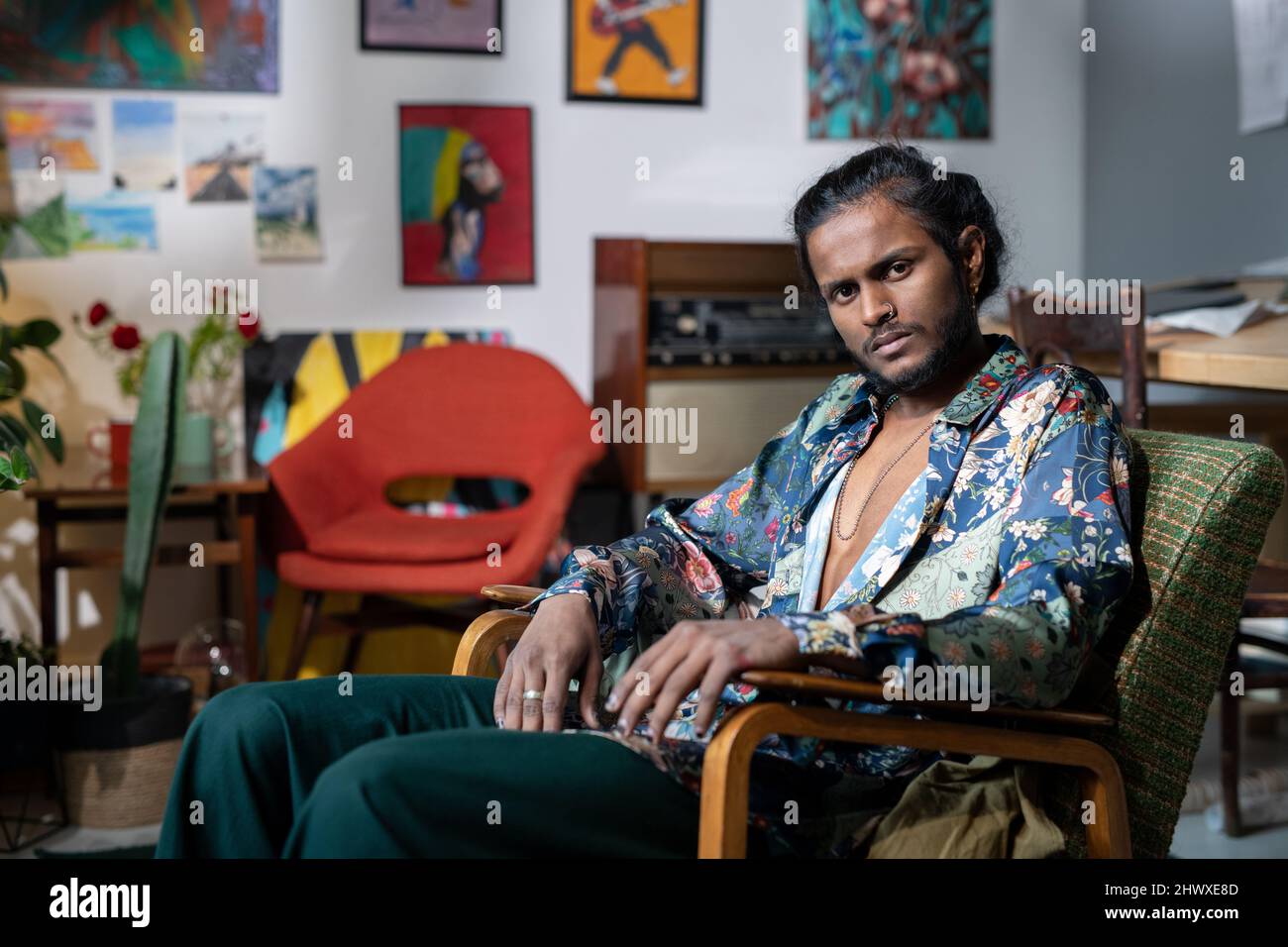 Junger seriöser, nicht-binärer Mann in trendigen Kleidern sitzt im Sessel vor der Kamera und ruht sich im Studio für moderne Kunst aus Stockfoto