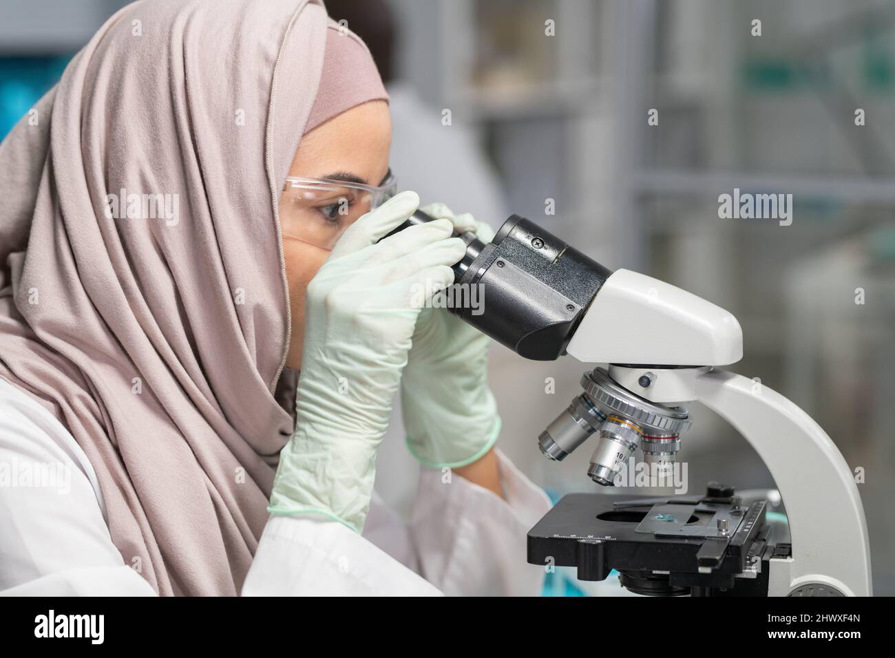 Seitenansicht einer jungen muslimischen Wissenschaftlerin in Hijab, Handschuhen und Schutzbrille, die neue chemische Verbindungen im Labor untersucht Stockfoto